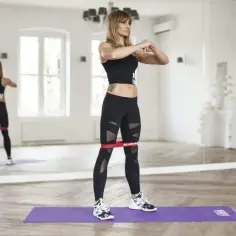 Kobieta wykonuje ćwiczenia z gumami mini band, wzmacniając nogi. Widoczna jest jej sylwetka w lustrzanym odbiciu.