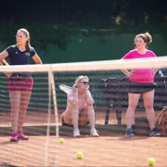 Trzy kobiety na korcie tenisowym, jedna stoi z rakietą, druga siedzi, trzecia odpoczywa.