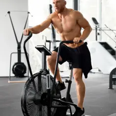 Mężczyzna z brodą intensywnie ćwiczy cardio na rowerze stacjonarnym w siłowni.