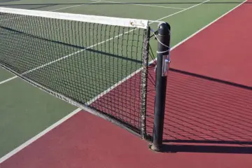 Siatka tenisowa na korcie, jej standardowa wysokosc siatki tenis jest kluczowa dla gry. Cień siatki pada na czerwoną nawierzchnię.